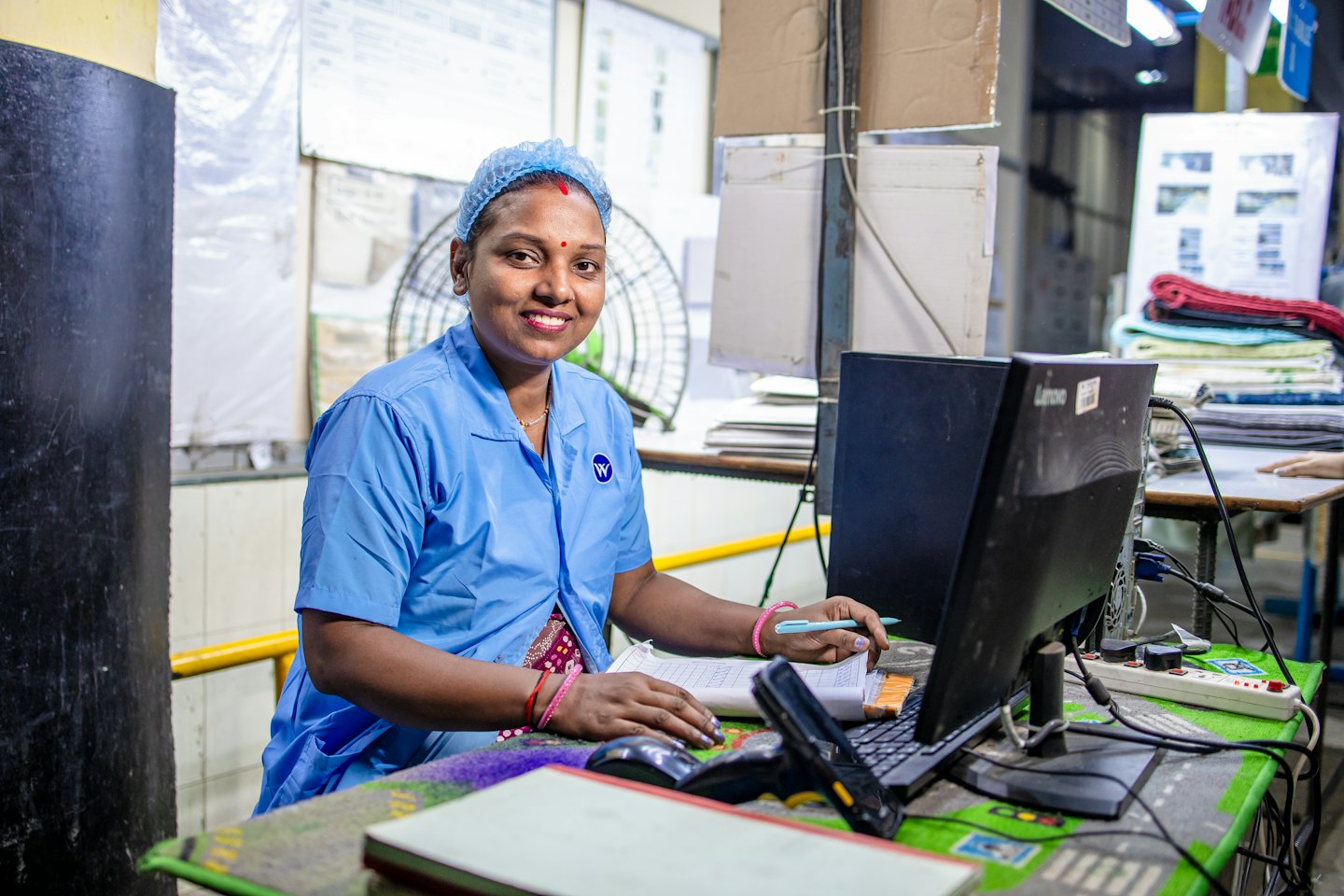 Smiling woman in blue uniform works at a busy factory desk.