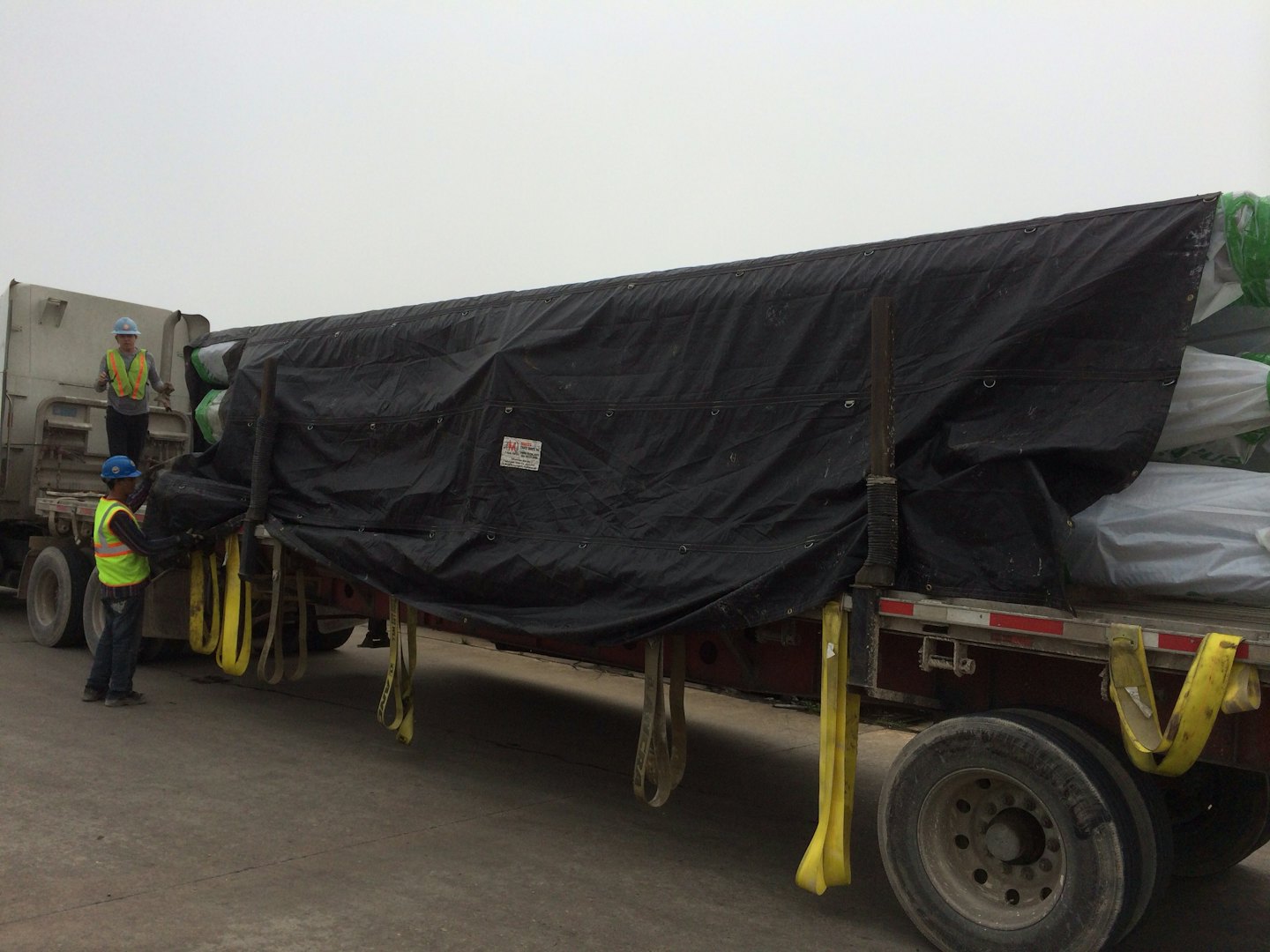 Workers securing a tarp-covered load of pipes on a semi-truck.